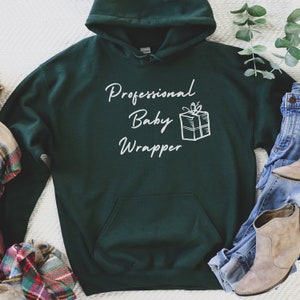 Puede incluir: Una sudadera con capucha verde oscuro con el texto "Professional Baby Wrapper" en letras blancas y un contorno blanco de una caja de regalo con un lazo en la parte superior.