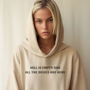 May include: A beige hoodie with a front pocket and a hood. The text "HELL IS EMPTY AND ALL THE DEVILS ARE HERE" is printed in black on the front. The hoodie is made of a soft material.