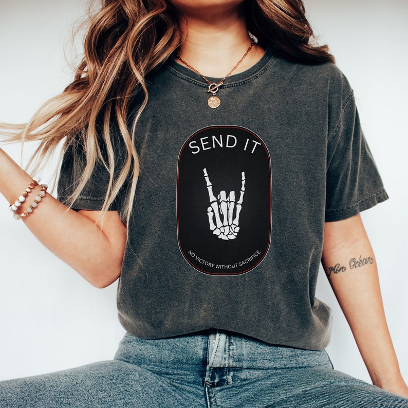 May include: A dark gray t-shirt with a black oval graphic featuring a white skeleton hand making the rock and roll sign. The graphic also includes the text "SEND IT" and "NO VICTORY WITHOUT SACRIFICE".