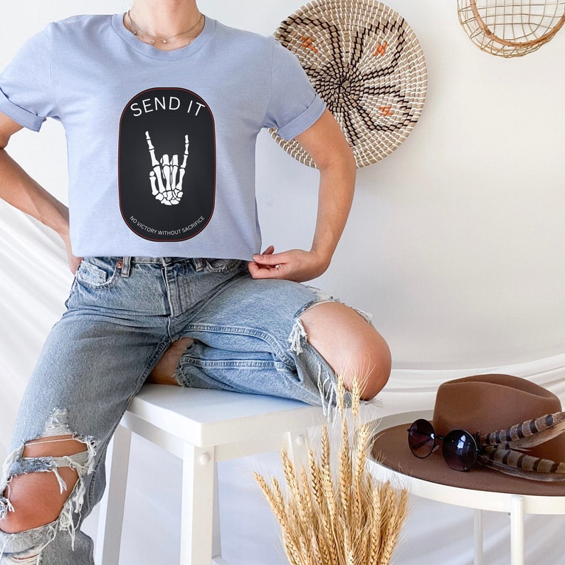May include: A light blue t-shirt with a black oval graphic that says "Send It" and features a white skeleton hand making a rock and roll sign. The text below the hand says "No victory without sacrifice."