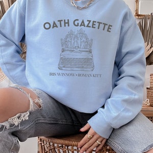 Puede incluir: Una sudadera azul claro con el texto "OATH GAZETTE" impreso en gris. La sudadera presenta un gr&aacute;fico de una m&aacute;quina de escribir con flores y el texto "IRIS WINNOW - ROMAN KITT" impreso debajo del gr&aacute;fico.