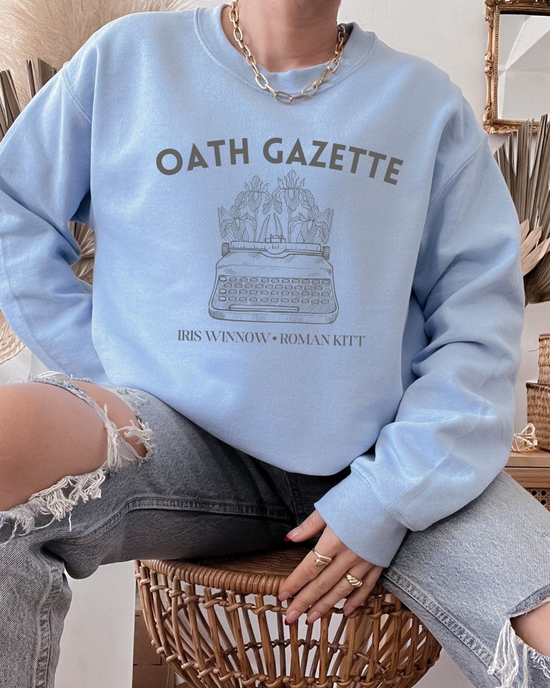 Puede incluir: Una sudadera azul claro con el texto "OATH GAZETTE" impreso en gris. La sudadera presenta un gr&aacute;fico de una m&aacute;quina de escribir con flores y el texto "IRIS WINNOW - ROMAN KITT" impreso debajo del gr&aacute;fico.