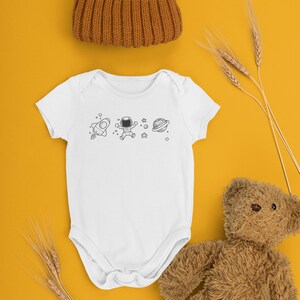 May include: A white baby onesie with a black and white astronaut, rocket, and planet design. The onesie is on a yellow background with a brown teddy bear and wheat stalks.