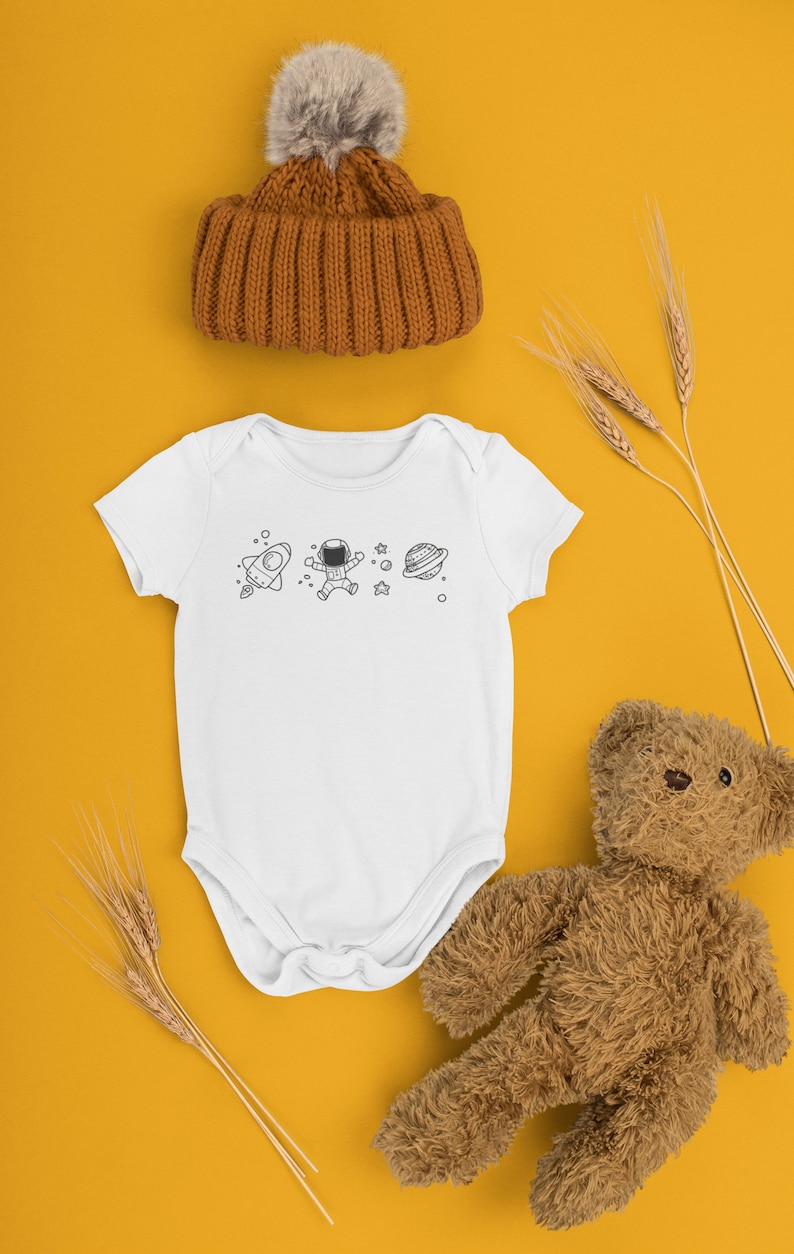 May include: A white baby onesie with a black and white astronaut, rocket, and planet design. The onesie is on a yellow background with a brown teddy bear and wheat stalks.