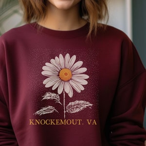 Puede incluir: Sudadera color burdeos con un gr&aacute;fico de margarita blanca y el texto "Knockemout, VA" en naranja.
