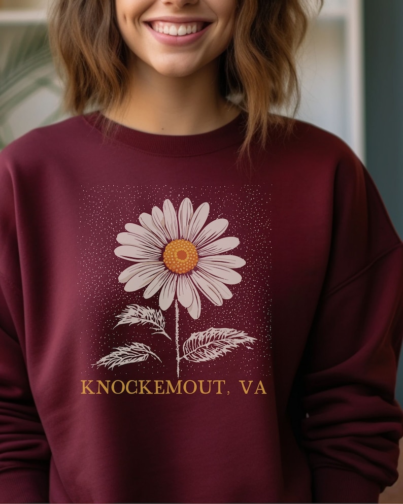 Puede incluir: Sudadera color burdeos con un gr&aacute;fico de margarita blanca y el texto "Knockemout, VA" en naranja.