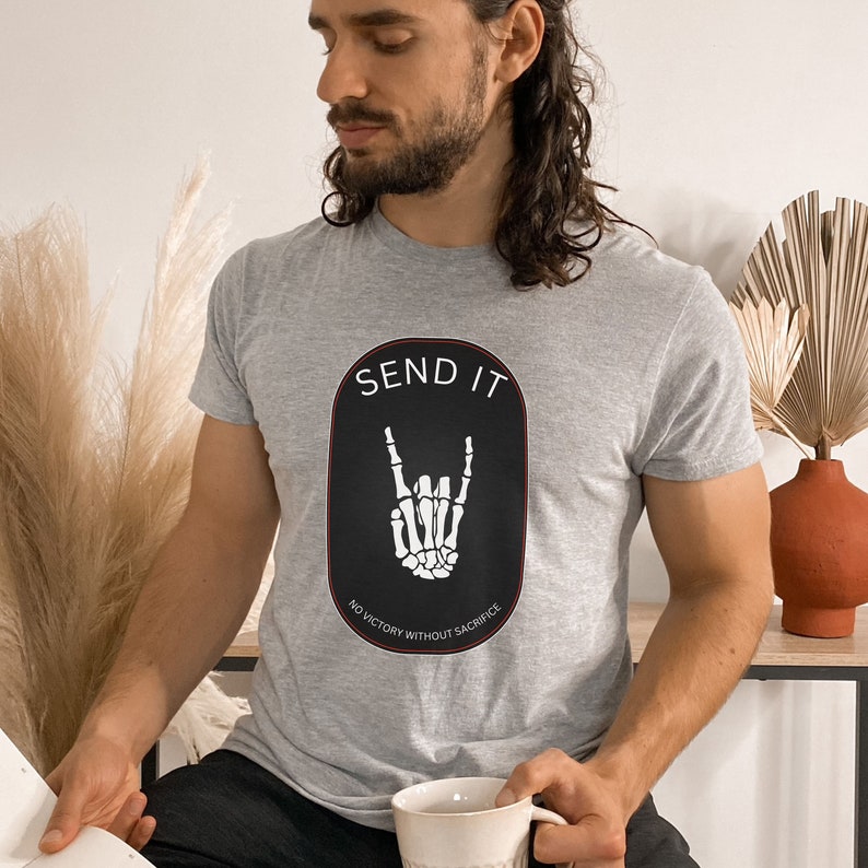 May include: A gray t-shirt with a black oval graphic featuring a white skeleton hand making a rock and roll sign. The graphic also includes the text "SEND IT" and "NO VICTORY WITHOUT SACRIFICE".