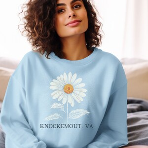 Puede incluir: Una sudadera azul claro con un gr&aacute;fico de margarita blanca y el texto "Knockemout, VA".