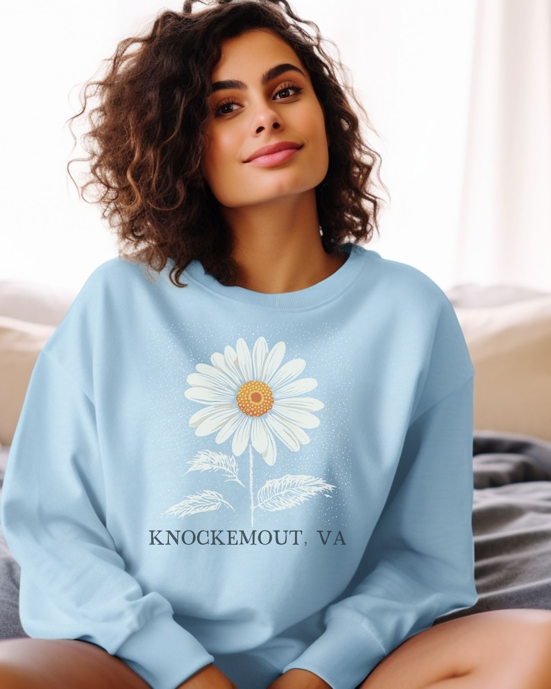 Puede incluir: Una sudadera azul claro con un gr&aacute;fico de margarita blanca y el texto "Knockemout, VA".
