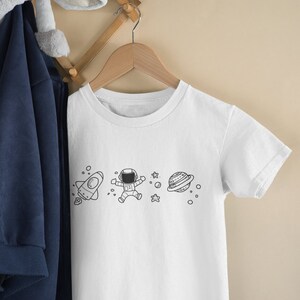 May include: White t-shirt with a black and white graphic of an astronaut, a rocket, a planet, and stars.
