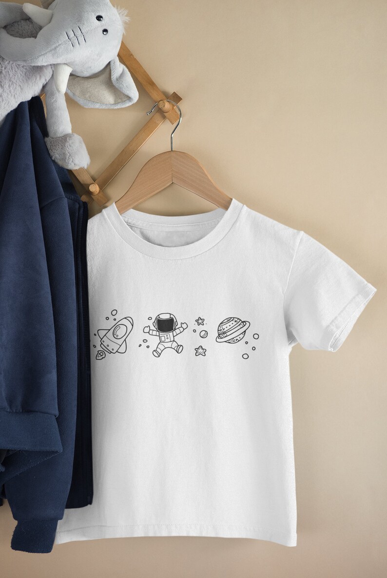 May include: White t-shirt with a black and white graphic of an astronaut, a rocket, a planet, and stars.
