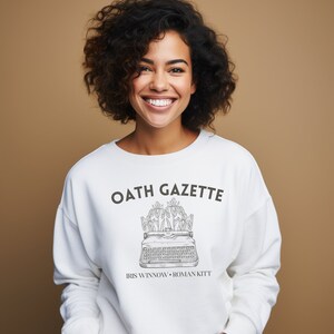 Puede incluir: Sudadera blanca con el texto "OATH GAZETTE" y un gr&aacute;fico de una m&aacute;quina de escribir con flores en la parte superior. El texto "IRIS WINNOW - ROMAN KITT" est&aacute; debajo del gr&aacute;fico.