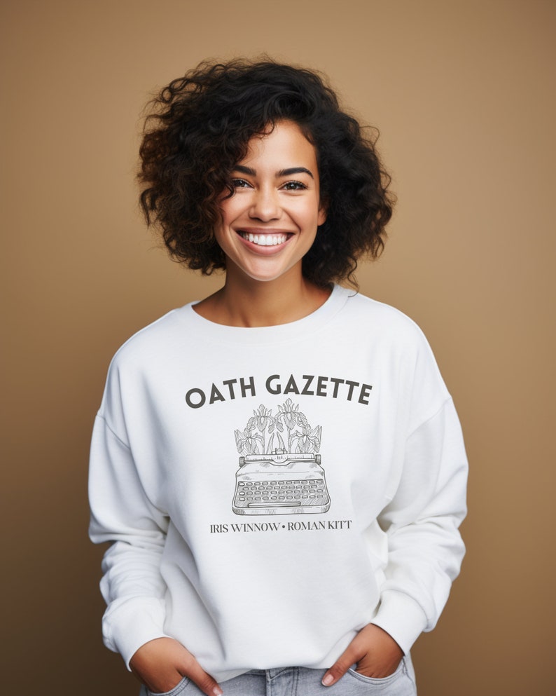 Puede incluir: Sudadera blanca con el texto "OATH GAZETTE" y un gr&aacute;fico de una m&aacute;quina de escribir con flores en la parte superior. El texto "IRIS WINNOW - ROMAN KITT" est&aacute; debajo del gr&aacute;fico.