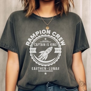 May include: A dark gray t-shirt with a white graphic design of a rocket ship and the text "Rampion Crew, Captain is King, Est. 126 T.E., Earthen Lunar".