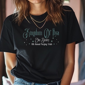 Może przedstawiać: Czarny t-shirt z turkusowym napisem "Kingdom Of Ilya", grafiką miecza oraz "Elite Society" i "6th Annual Purging Trials". Koszulka ma okrągły dekolt i krótkie rękawy.