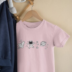 May include: A pink t-shirt with a white graphic of a rocket, astronaut, stars, and a planet. The t-shirt is hanging on a wooden hanger.