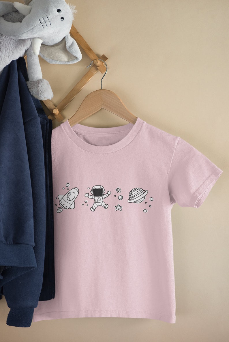 May include: A pink t-shirt with a white graphic of a rocket, astronaut, stars, and a planet. The t-shirt is hanging on a wooden hanger.