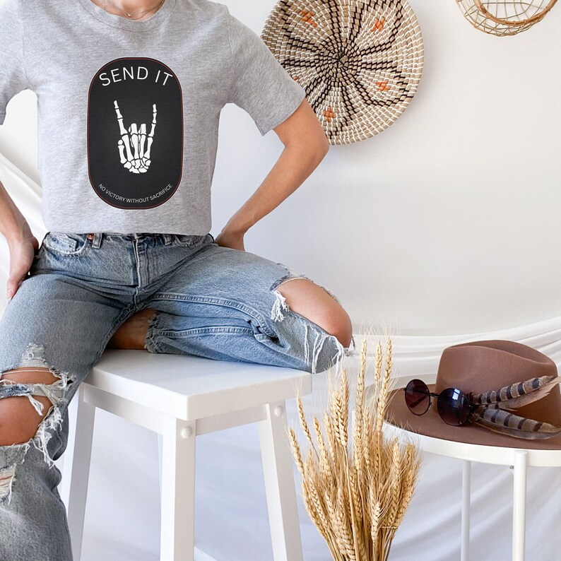 May include: A gray cropped t-shirt with a black and white graphic of a skeleton hand making a rock and roll sign. The text "SEND IT" is above the graphic and "NO VICTORY WITHOUT SACRIFICE" is below the graphic. The shirt is being worn by a person sitting on a white stool.