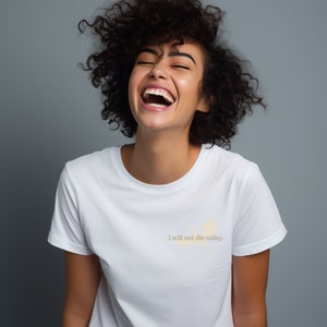 May include: A woman with curly brown hair laughs while wearing a white t-shirt with the text "I will not die today." printed on it.