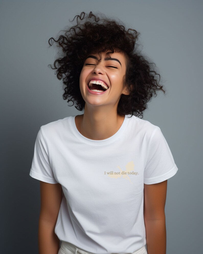 May include: A woman with curly brown hair laughs while wearing a white t-shirt with the text "I will not die today." printed on it.