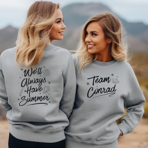 Peut inclure: Deux femmes portent des sweat-shirts gris avec du texte noir. L'un des sweat-shirts dit "We'll Always Have Summer" avec des graphiques de coquillages. L'autre dit "Team Conrad" avec un graphique de coquillage. L'arrière-plan est une scène extérieure floue.