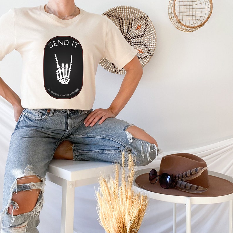 May include: A woman wearing a cream colored t-shirt with a black oval graphic that says "SEND IT" and "NO VICTORY WITHOUT SACRIFICE" with a skeleton hand making a rock and roll sign. She is wearing blue jeans with rips in the knees and is sitting on a white table.