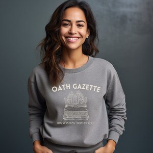 Puede incluir: Sudadera gris con un gr&aacute;fico blanco de una m&aacute;quina de escribir con flores y el texto "OATH GAZETTE" e "IRIS WINNOW - ROMAN KITT"