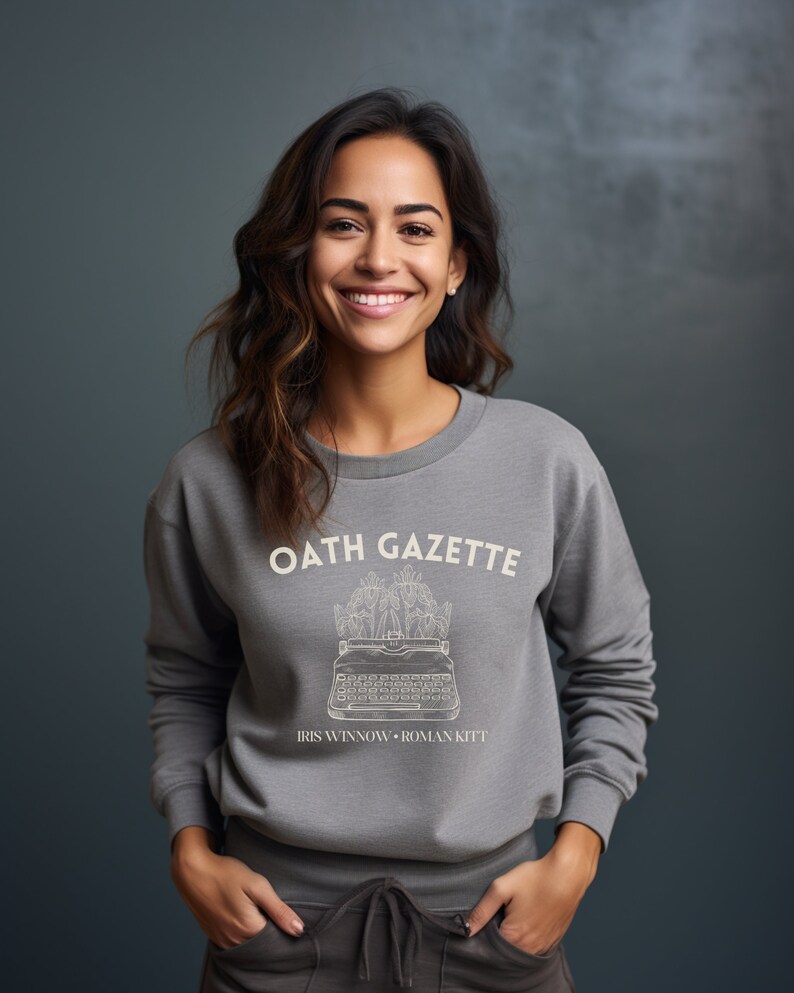 Puede incluir: Sudadera gris con un gr&aacute;fico blanco de una m&aacute;quina de escribir con flores y el texto "OATH GAZETTE" e "IRIS WINNOW - ROMAN KITT"