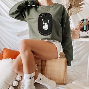 May include: A green cropped sweatshirt with the text "SEND IT" and a black oval graphic with a skeleton hand making a rock and roll sign. The text below the graphic reads "NO VICTORY WITHOUT SACRIFICE."