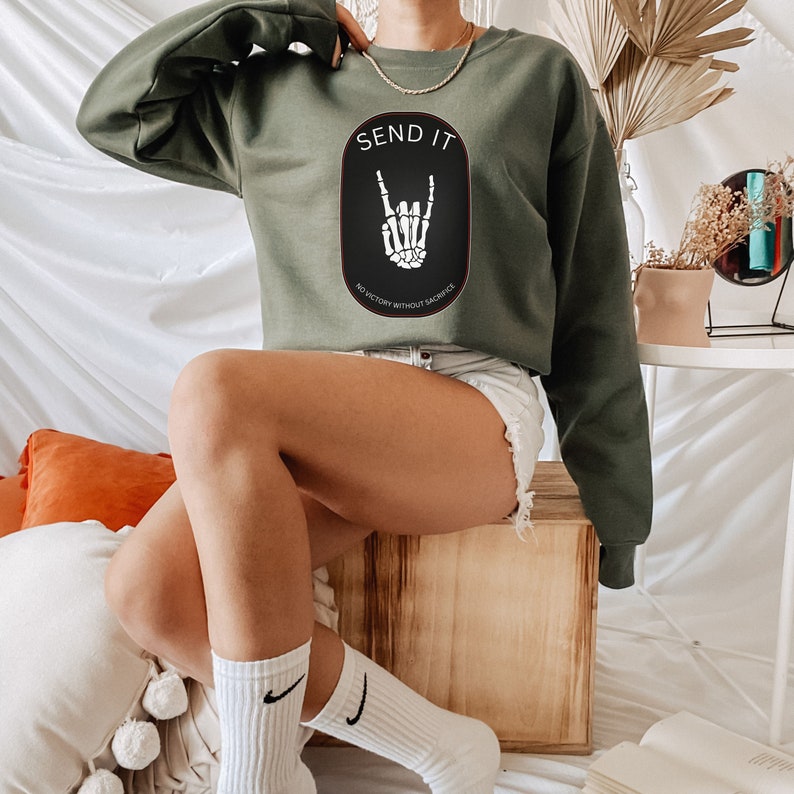 May include: A green cropped sweatshirt with the text "SEND IT" and a black oval graphic with a skeleton hand making a rock and roll sign. The text below the graphic reads "NO VICTORY WITHOUT SACRIFICE."