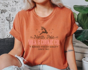 May include: A woman wearing a burnt orange t-shirt with a brown reindeer graphic and the text "North Pole Milk & Cookie Co.  > Baked Fresh Daily <  . Since 1842."
