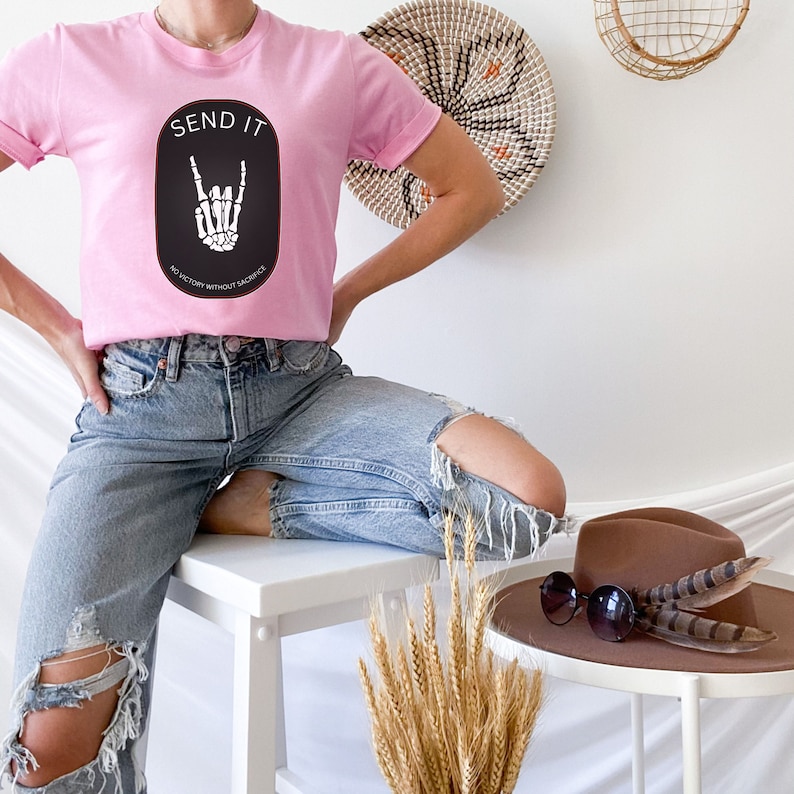 May include: A pink t-shirt with a black and white graphic of a skeleton hand making a rock and roll sign. The graphic is surrounded by a black circle with the text "SEND IT" and "NO VICTORY WITHOUT SACRIFICE" in white.