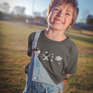 May include: A young person wearing a dark grey t-shirt with a white astronaut, planet, and stars design. The t-shirt is worn over a pair of blue denim overalls.