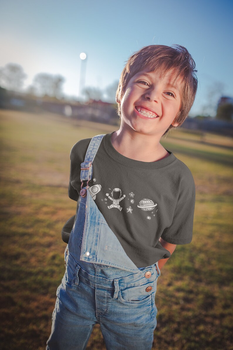 May include: A young person wearing a dark grey t-shirt with a white astronaut, planet, and stars design. The t-shirt is worn over a pair of blue denim overalls.