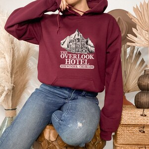 May include: A maroon hoodie with a white graphic of a mountain and a building with the text "The Overlook Hotel Sidewinder, Colorado".