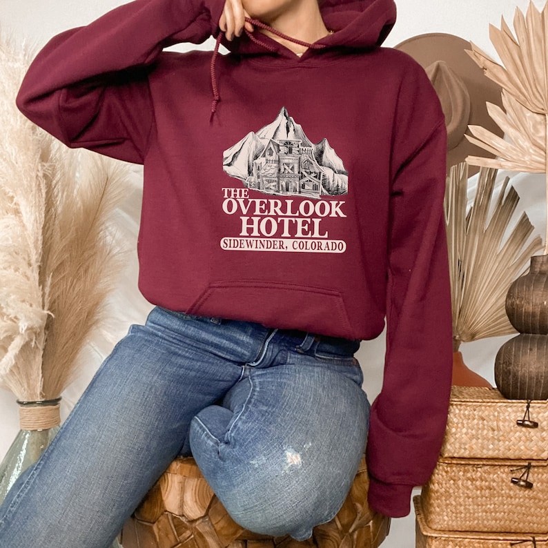 May include: A maroon hoodie with a white graphic of a mountain and a building with the text "The Overlook Hotel Sidewinder, Colorado".