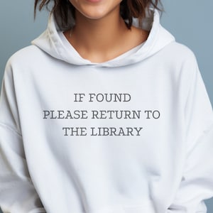 May include: White hooded sweatshirt with the text "IF FOUND PLEASE RETURN TO THE LIBRARY" in gray. The sweatshirt has a drawstring hood and a front pocket. The person wearing the sweatshirt is smiling.