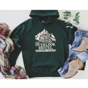 May include: A dark green hooded sweatshirt with a white graphic print of the Overlook Hotel from the movie "The Shining". The graphic includes the text "The Overlook Hotel" and "Sidewinder, Colorado".