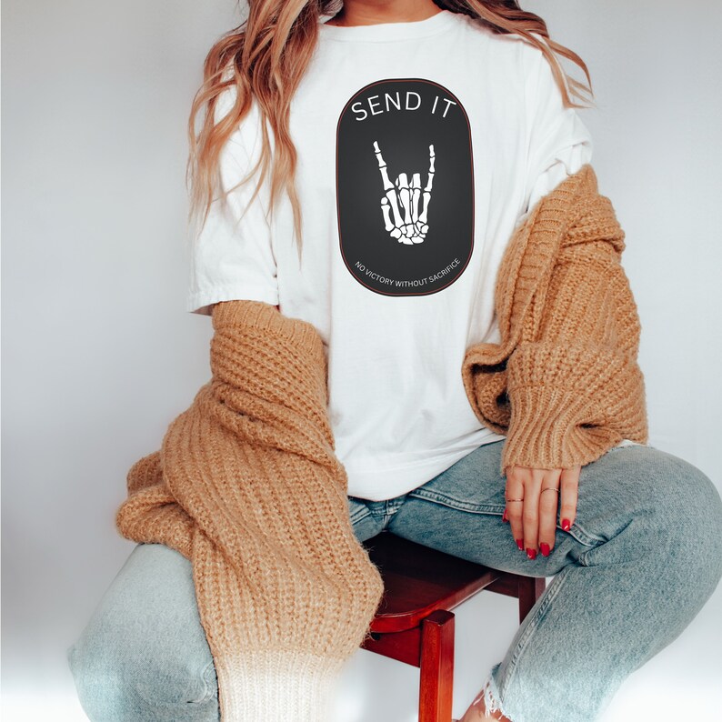 May include: White t-shirt with a black oval graphic featuring a skeleton hand making a rock and roll sign. The text "SEND IT" is above the hand and "NO VICTORY WITHOUT SACRIFICE" is below the hand.