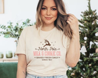 May include: A beige t-shirt with a brown reindeer graphic and the text "North Pole Milk & Cookie Co." in brown and pink. The text "Baked Fresh Daily" and "Since 1842" are in pink.
