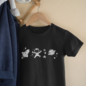 May include: A black t-shirt with a white astronaut, rocket, planet, and stars graphic. The t-shirt is hanging on a wooden hanger.