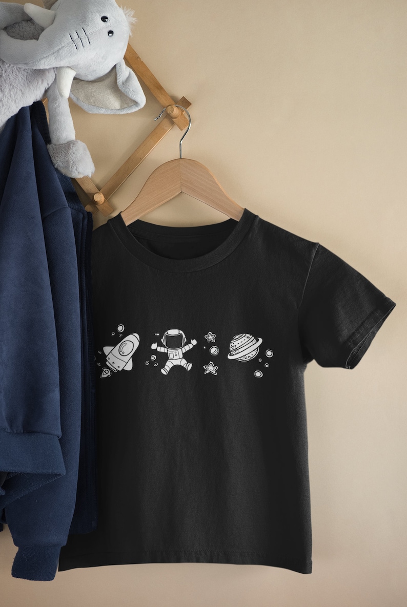 May include: A black t-shirt with a white astronaut, rocket, planet, and stars graphic. The t-shirt is hanging on a wooden hanger.