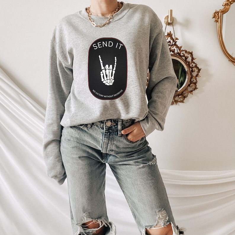 May include: Gray sweatshirt with a black and white graphic of a skeleton hand making a rock and roll sign. The graphic is inside a black oval with the text "SEND IT" in white above the hand and "NO VICTORY WITHOUT SACRIFICE" in white below the hand.
