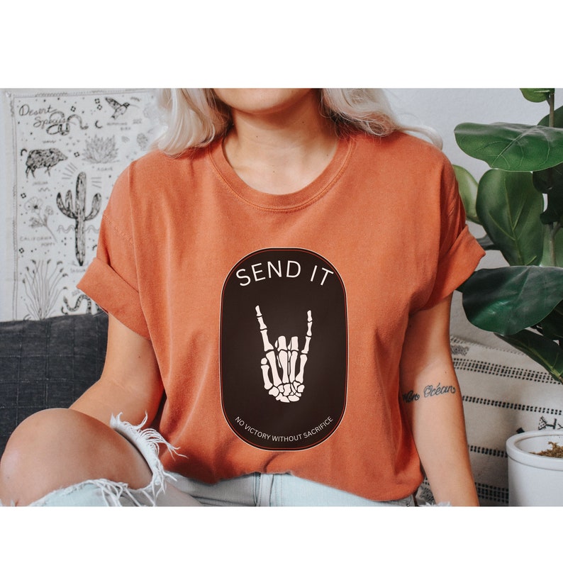 May include: A burnt orange t-shirt with a black oval graphic that says "SEND IT" in white text. The graphic also features a skeleton hand making the rock and roll hand gesture. Below the hand is the text "NO VICTORY WITHOUT SACRIFICE" in white text.