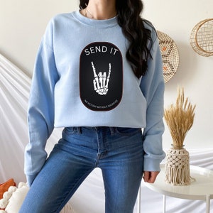 May include: Light blue cropped sweatshirt with a black oval graphic featuring a skeleton hand making a rock and roll sign. The graphic also includes the text "SEND IT" and "NO VICTORY WITHOUT SACRIFICE".