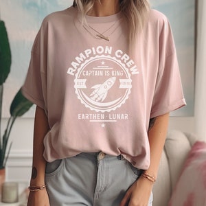 May include: A light pink t-shirt with a white graphic print of a rocket ship and the text "Rampion Crew, Captain is King, Est. 126 TE, Earthen Lunar".