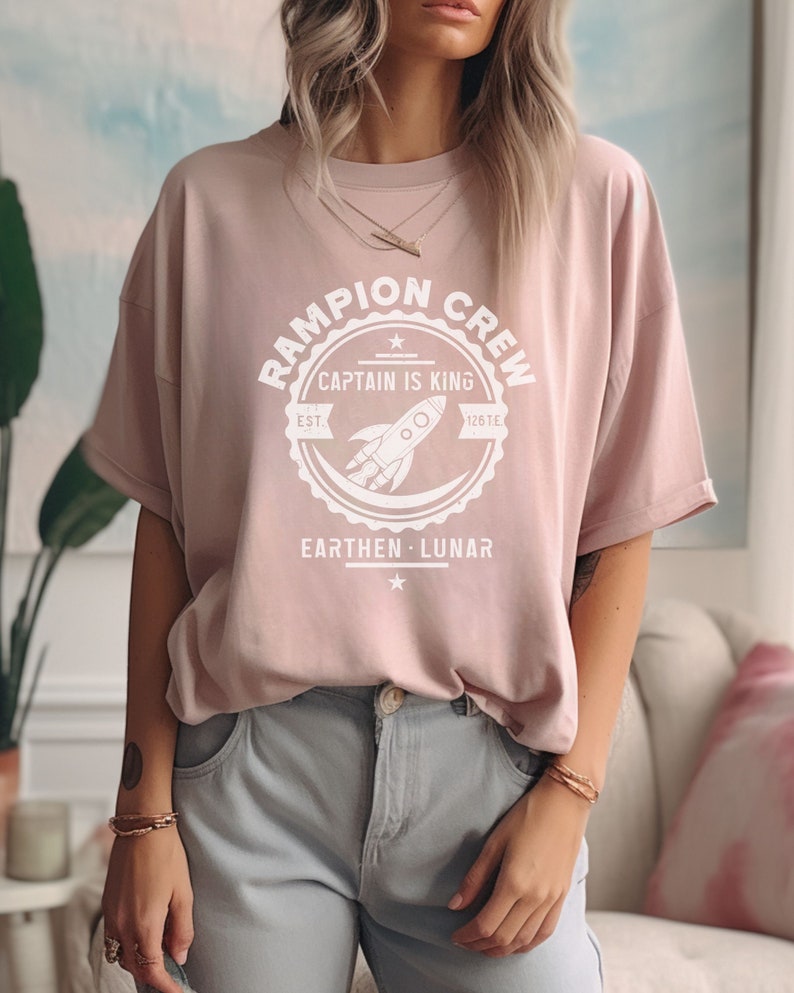May include: A light pink t-shirt with a white graphic print of a rocket ship and the text "Rampion Crew, Captain is King, Est. 126 TE, Earthen Lunar".
