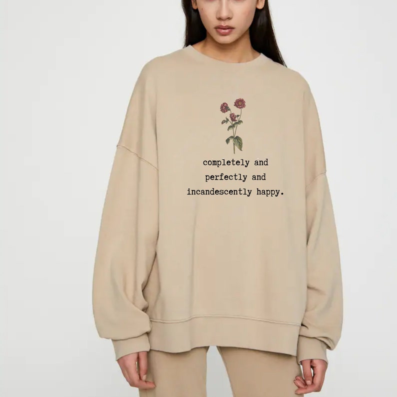 Puede incluir: Una sudadera beige con un gr&aacute;fico floral y el texto "completely and perfectly and incandescently happy."