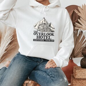 May include: White hoodie with a black graphic print of a mountain with a building on it. The text "The Overlook Hotel Sidewinder, Colorado" is printed below the graphic.