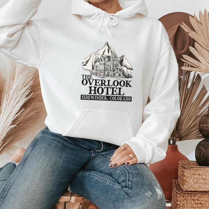 May include: White hoodie with a black graphic print of a mountain with a building on it. The text "The Overlook Hotel Sidewinder, Colorado" is printed below the graphic.